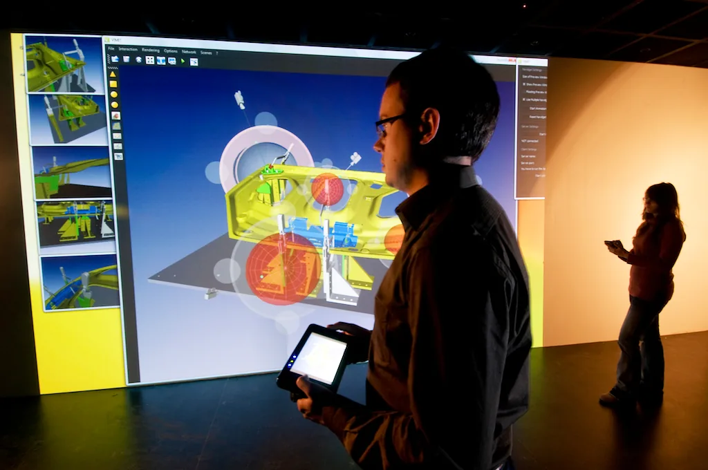 A researcher stands before a large stereo projection of a CAD assembly, driving the view from a handheld tablet.