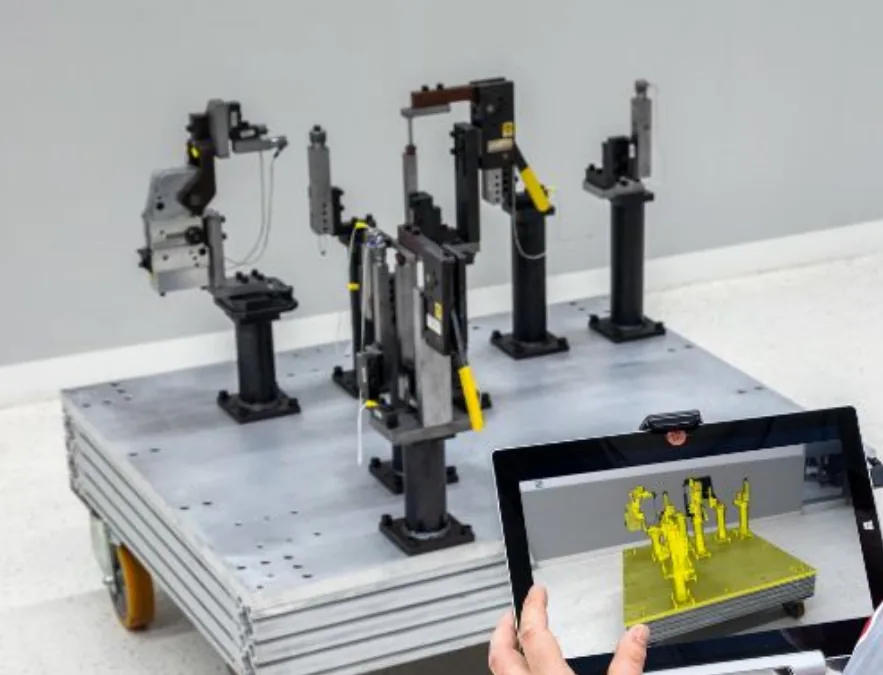 A tablet held in front of a metal fixture table shows the CAD model overlaid in yellow, aligned to the real hardware.